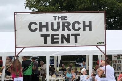 sign saying the church tent in front of tent with people in.png
